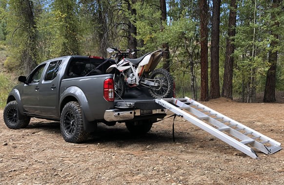A 5-step dirt bike ramp is shown in the image, leading up to the bed of a gray pickup truck. The ramp is made of rugged aluminum, and a white dirt bike is partially loaded onto the truck. The scene takes place in a wooded area with tall trees in the background.